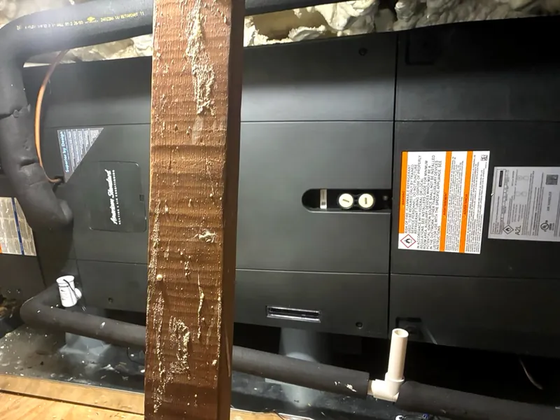 Black American Standard air handler on a platform in an attic, with insulated refrigerant lines and a condensate drain